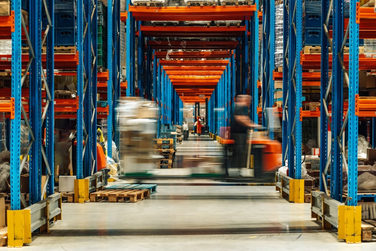 Warehouse operatives moving goods on forklifts between tall storage aisles